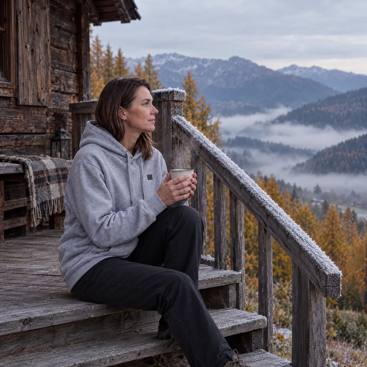 Frau im grauen beheizten Hoodie mit Tee auf der Hüttenterrasse am Herbstmorgen